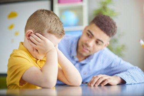 A child in a yellow shirt sits with his head in his hands, covering his eyes. A young adult in a blue shirt sits beside him, leaning toward him in a supportive manner.